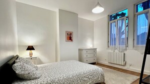 Ein Schlafzimmer mit Bett, Nachttisch, Lampe, Kommode, Heizkörper und Fenstern.
