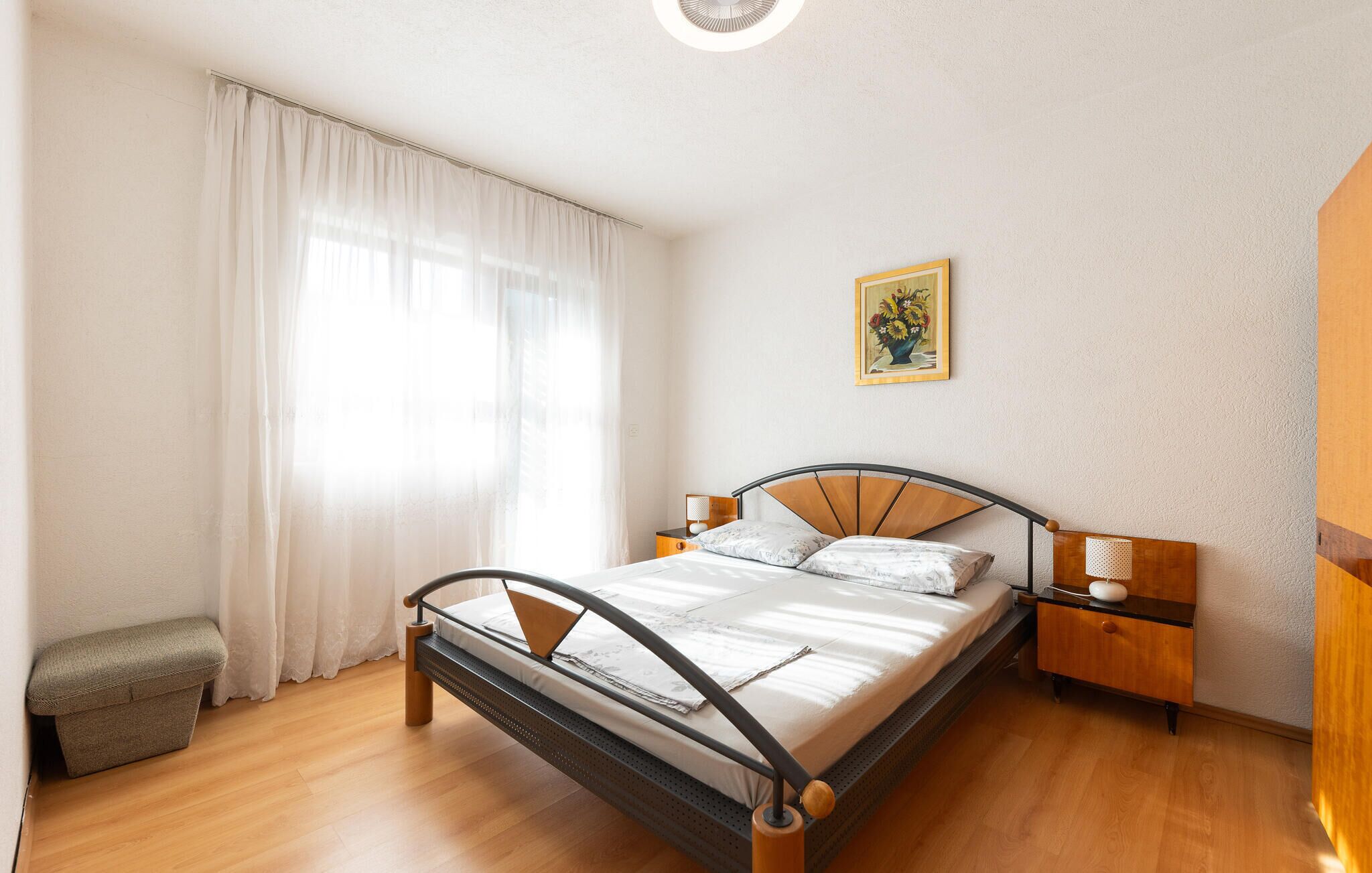 A bedroom with a bed, bedside tables, a nightstand, a small bench, a window with curtains, and a painting on the wall.