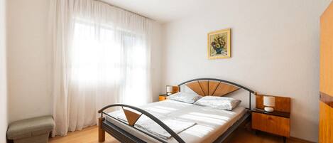 A bedroom with a bed, bedside tables, a nightstand, a small bench, a window with curtains, and a painting on the wall.