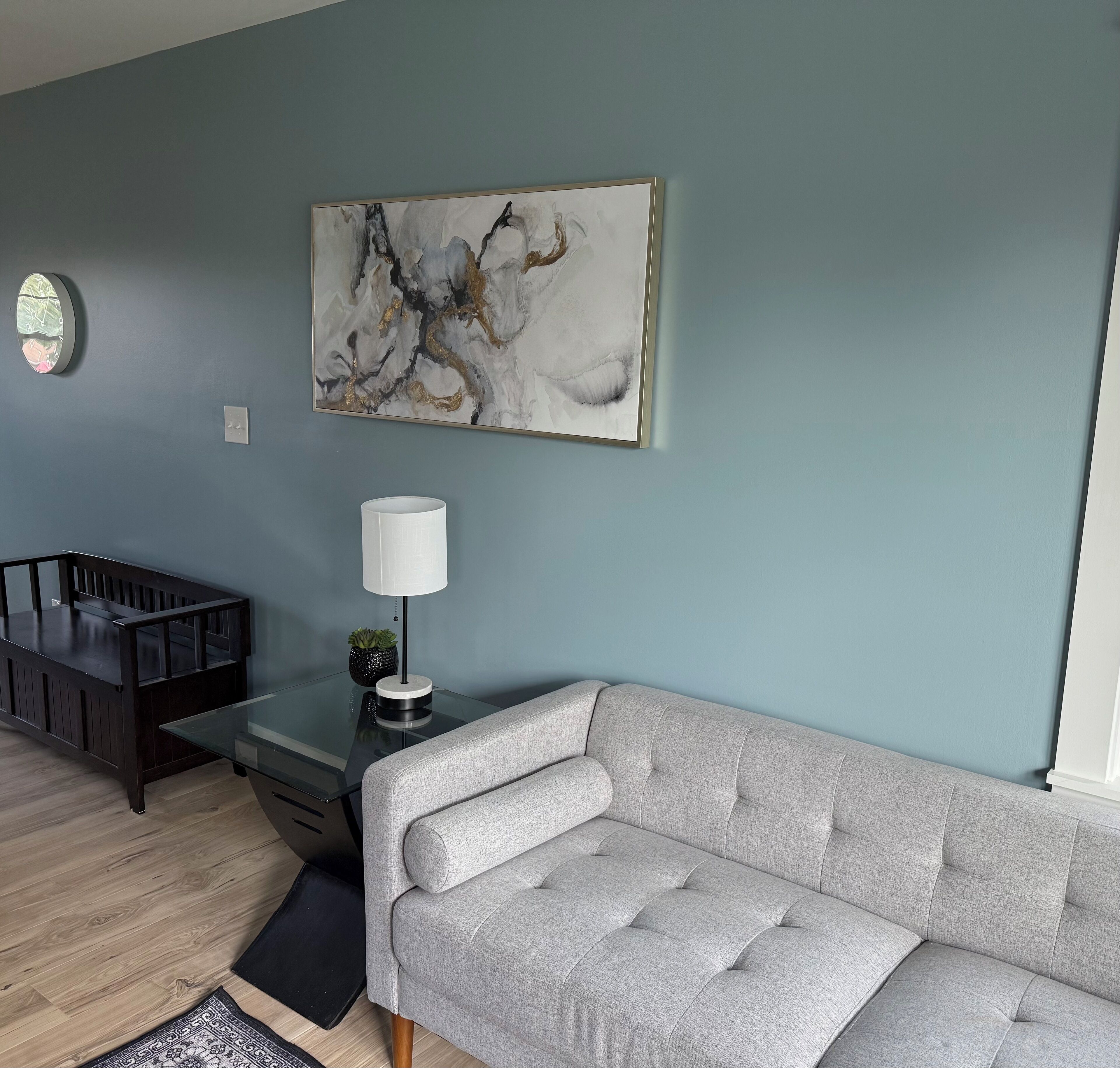 Un soggiorno moderno con un divano grigio, un tavolino da caffè in legno scuro, un tappeto rotondo, una lampada e un quadro astratto appeso al muro.