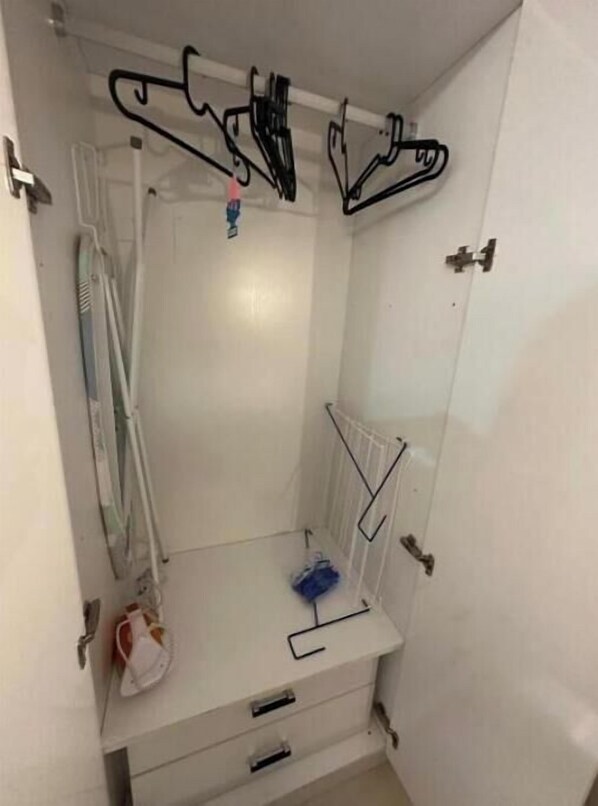 Un baño pequeño y blanco con ducha, inodoro y lavamanos. Hay varios perchas negras y un objeto azul sobre el lavamanos.