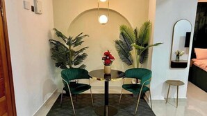 Un coin repas avec une table ronde, deux chaises vertes, un vase avec des fleurs rouges et deux grandes plantes en pot.