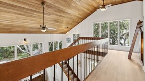 A spacious room with a high ceiling and wooden beams, a wooden staircase with a railing, and large windows offering a view of trees. - Charming Coastal Retreat sleeps up to 9 (Fernandina Beach)
