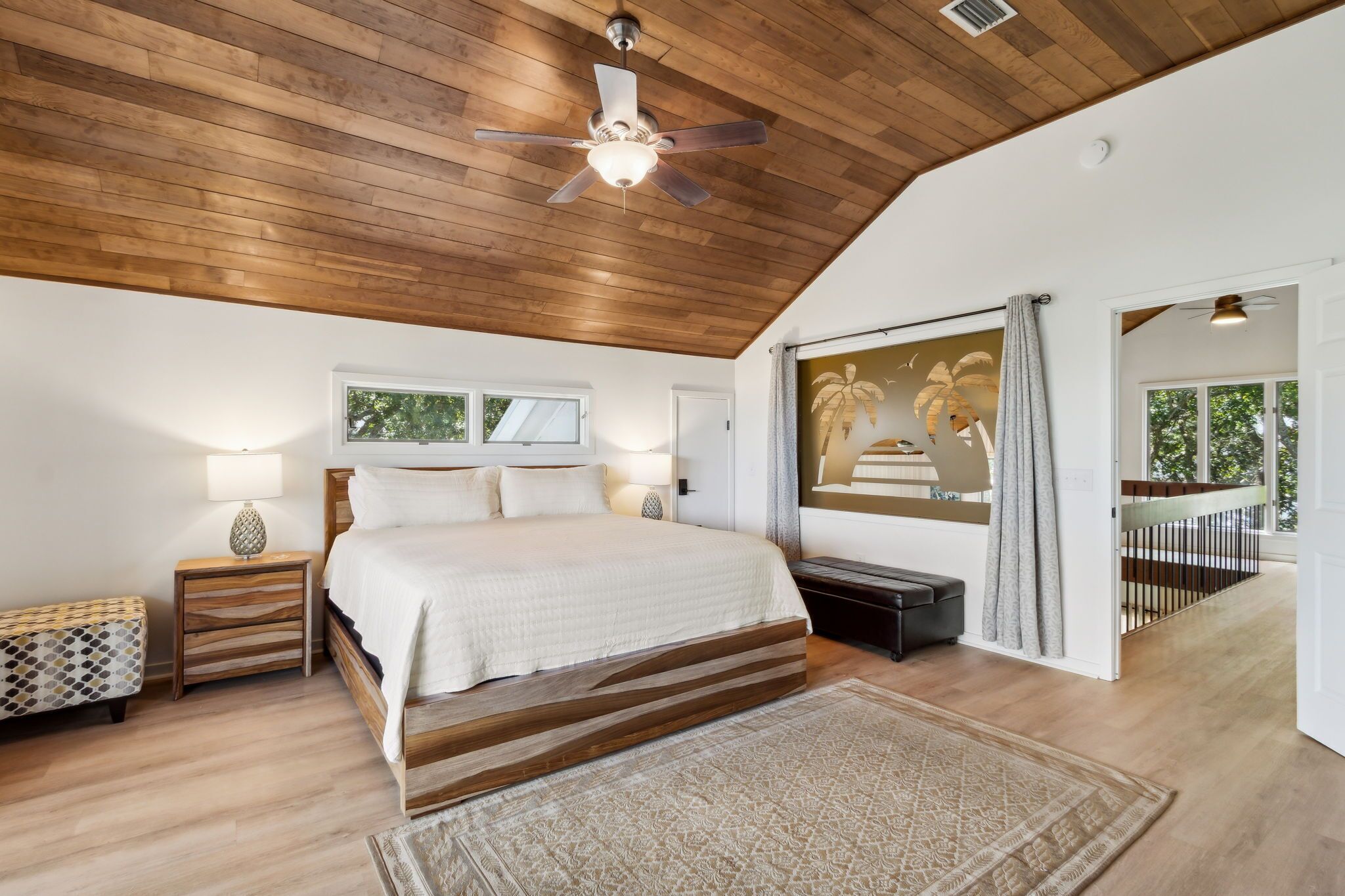 A bedroom with a wooden ceiling, a bed with white bedding, a nightstand with a lamp, a bench, and a large window with a view of palm trees.