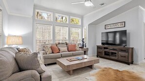 Un salon moderne avec un canapé, une table basse et un téléviseur. De grandes fenêtres sont équipées de stores, et on y trouve également un ventilateur de plafond ainsi qu’une enseigne indiquant « Hochatown, OK ».