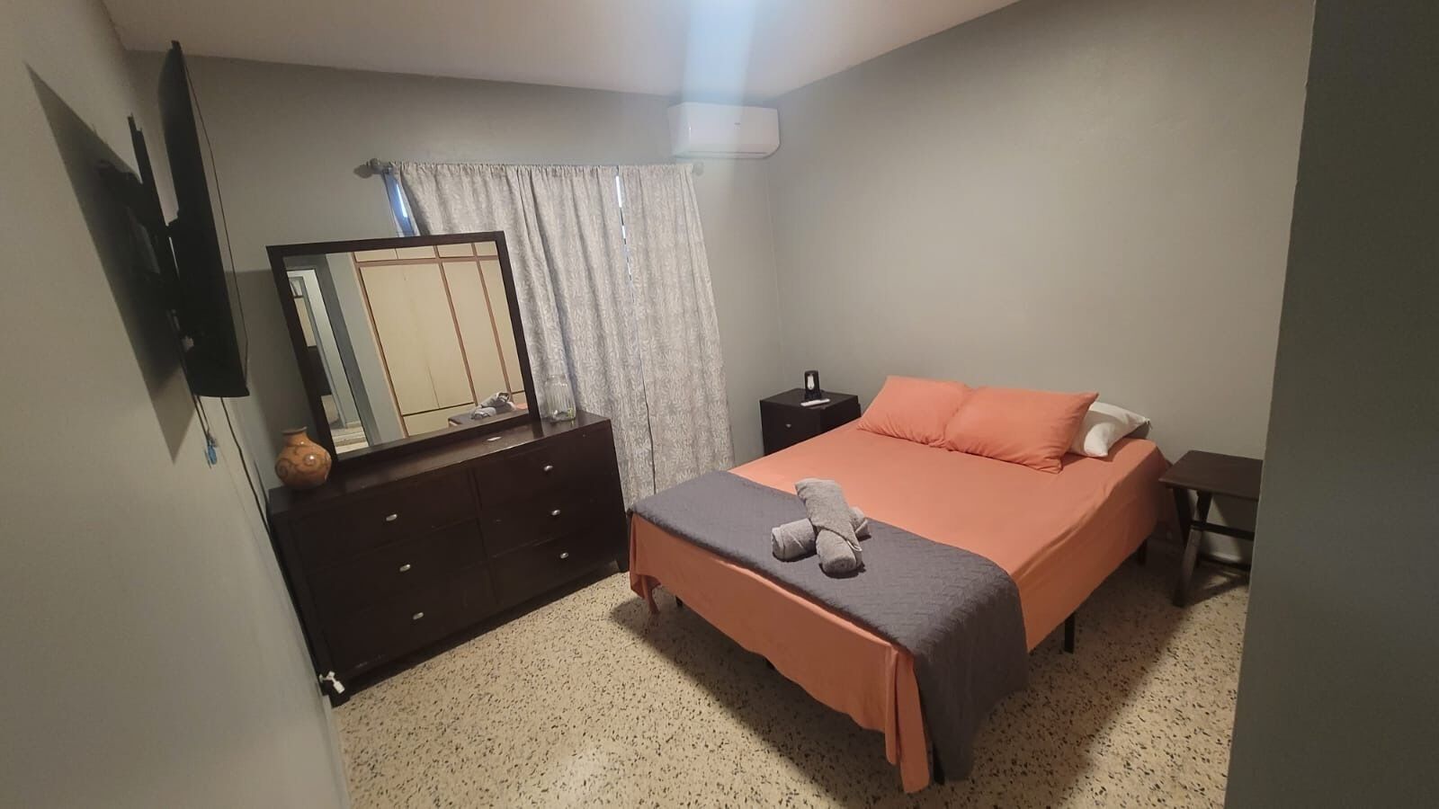 A bedroom with a bed, a dresser, a nightstand, a mirror, and a window with curtains.