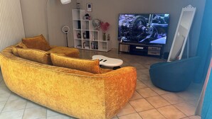 Een woonkamer met een ruime bank, een bijzettafeltje, een televisie, een boekenkast en een spiegel.