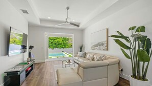 A modern living room with a beige sectional sofa, a large potted plant, and a view of a swimming pool through a glass door. - South Naples Pool House (Naples)