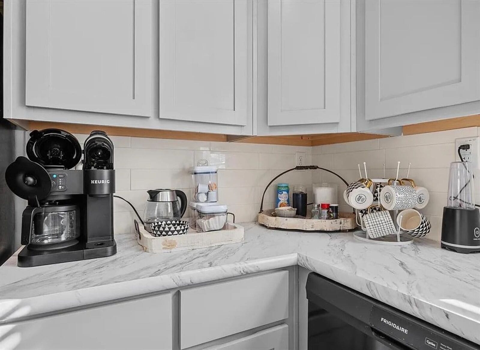 A kitchen with white cabinets, a coffee maker, a blender, and a dish rack with mugs.