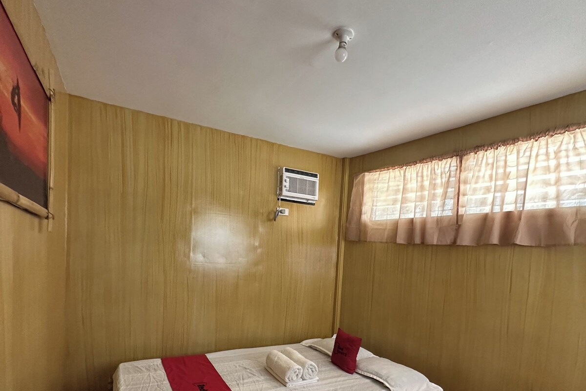 A single bed with a red blanket, a small table, and a window with curtains.