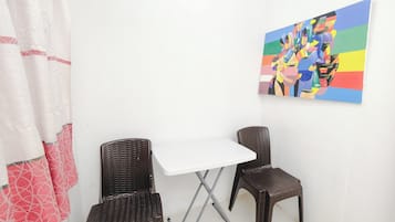 A small room with a white table and two wicker chairs, a colorful abstract painting on the wall, and a red and white patterned curtain.