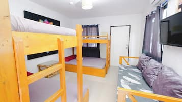 A dormitory room with bunk beds, a small table, a sofa, a TV, and an air conditioning unit.