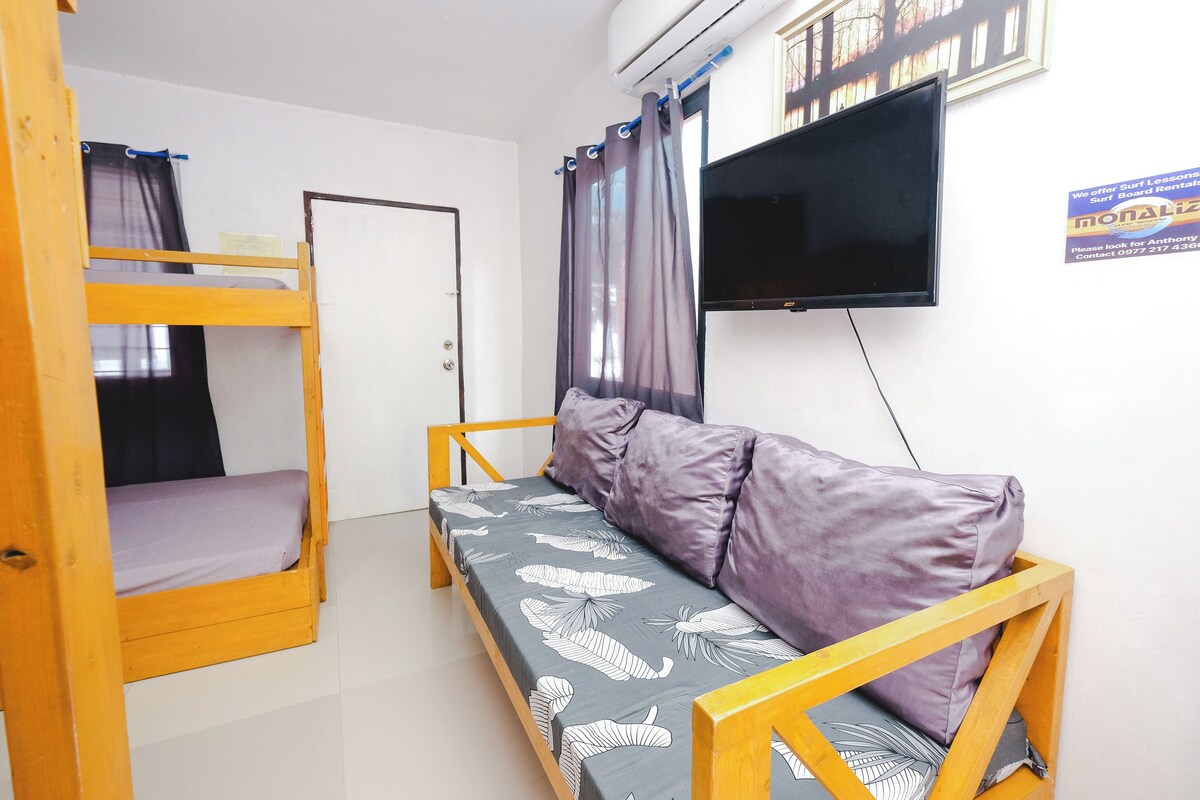 A room with bunk beds, a sofa, a flat-screen TV, and a sign about surf lessons.