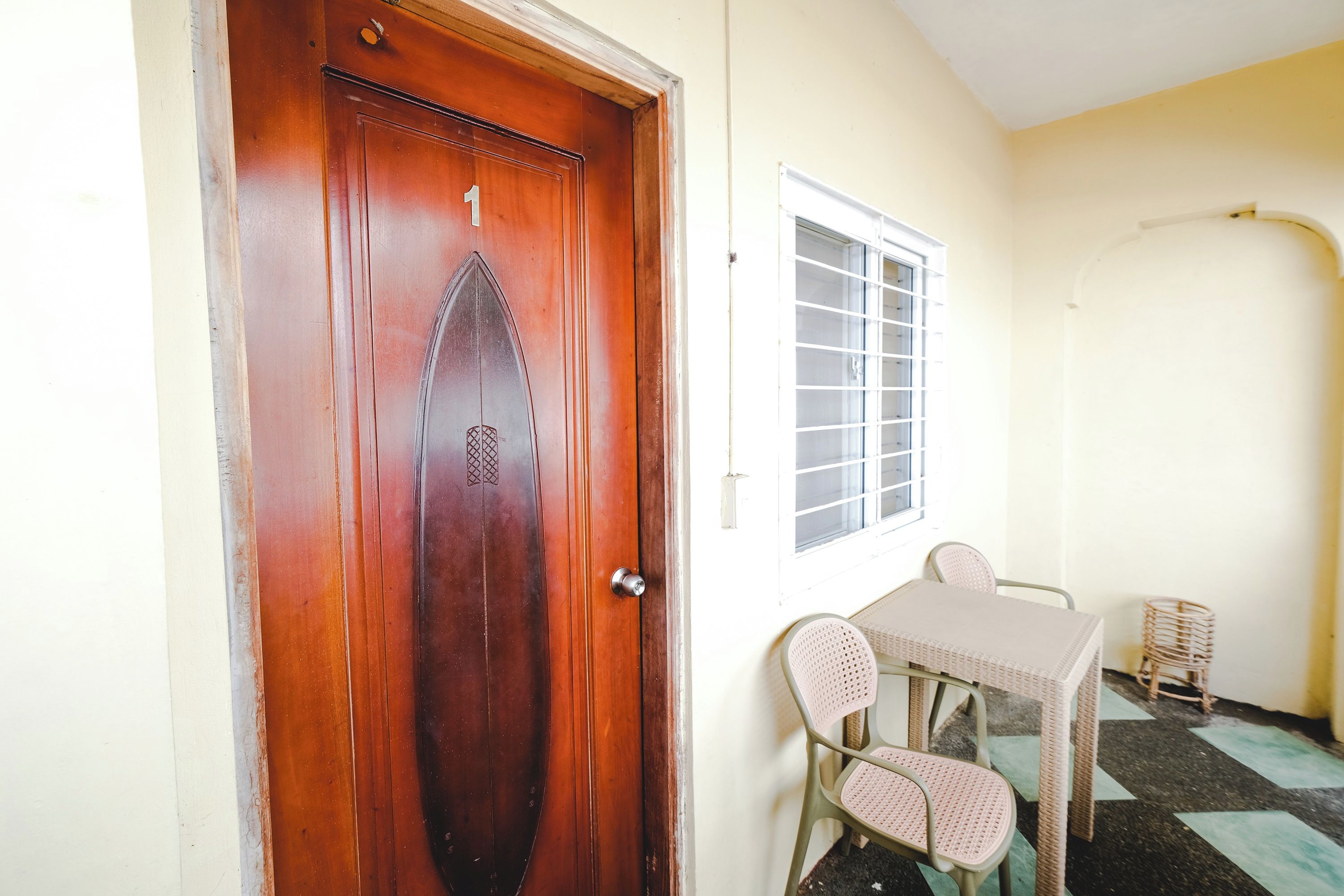 Una habitación con puerta de madera marcada con el número 1, ventana con barrotes, una mesa con dos sillas y una pequeña silla de mimbre.