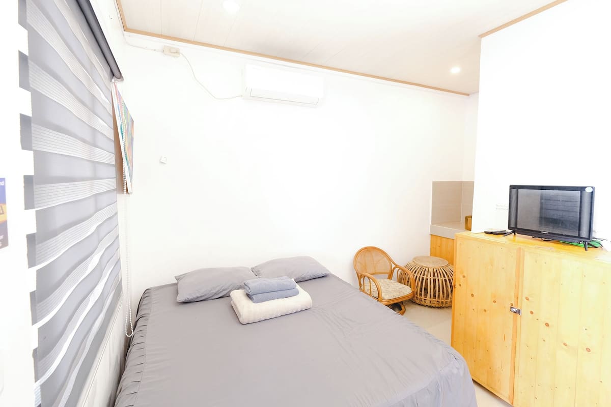 Un dormitorio con una cama, un televisor sobre un mueble de madera, una silla de mimbre y una pequeña mesa redonda.