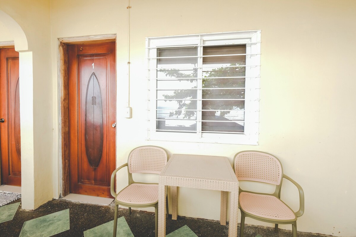 A room with a wooden door marked '1', a window with blinds, and two wicker chairs around a small table.