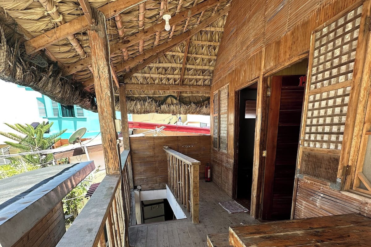 Una terraza de madera con techo de paja, barandillas de madera y vista a un edificio al fondo.
