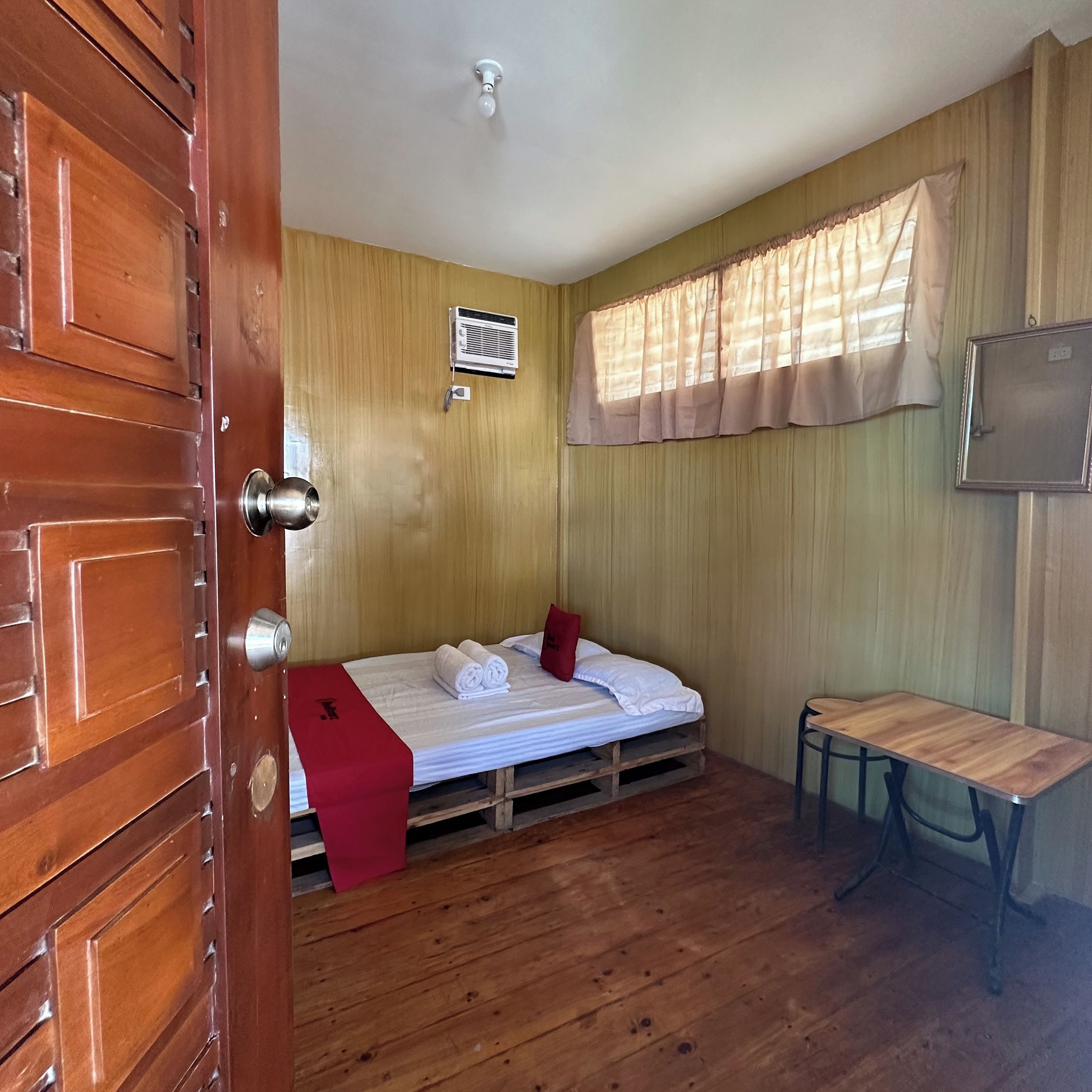 A compact room with a wooden door, a bed with white bedding, a small table, and a window with curtains.