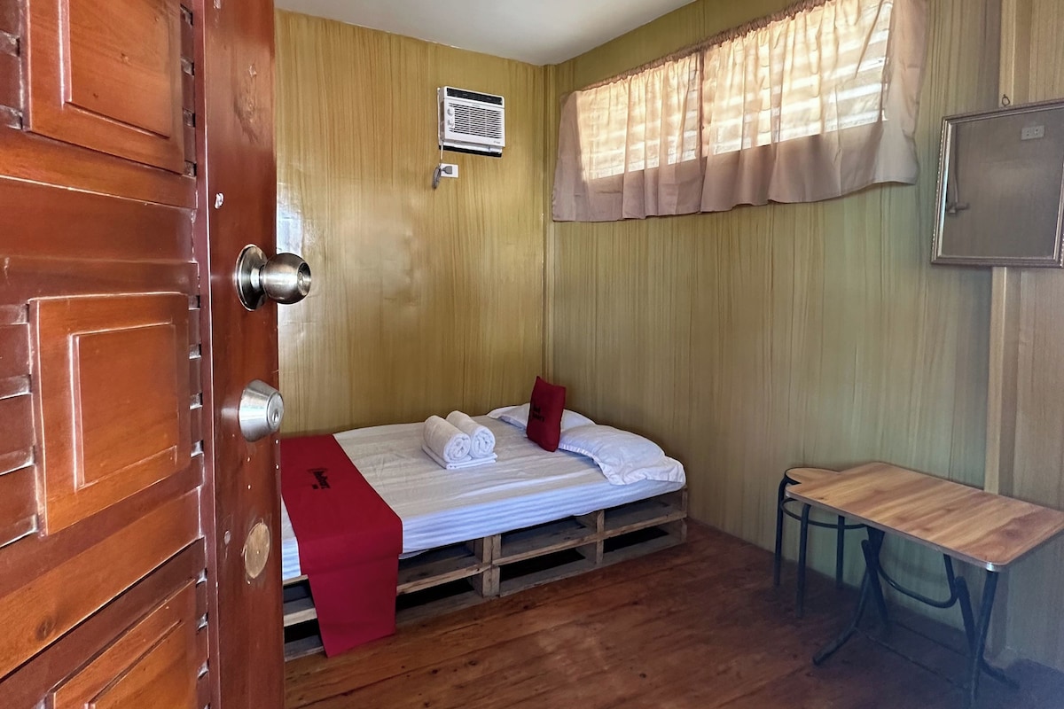 A compact room with a wooden door, a bed with white bedding, a small table, and a window with curtains.