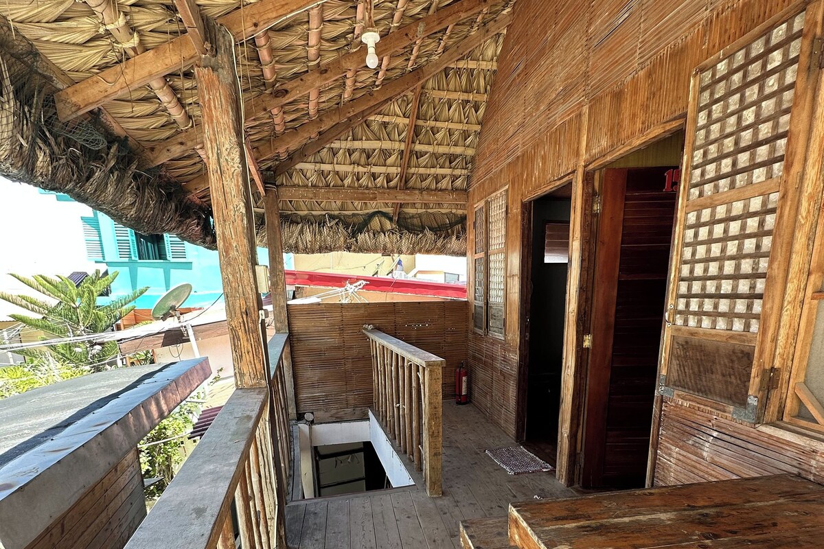 A wooden deck with a thatched roof, wooden railings, and a view of a building in the background.