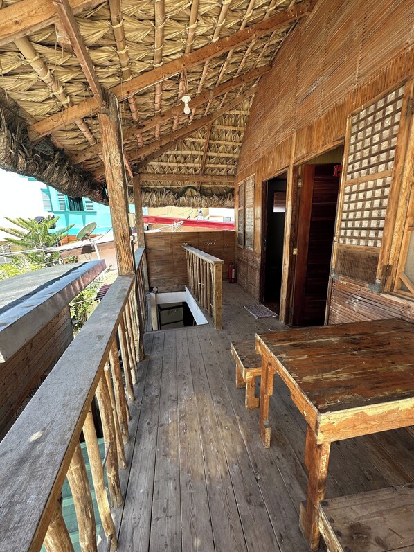 A wooden deck with a thatched roof, wooden railings, and a view of a building in the background. - Monaliza Surf Resort By Zuzu (San Juan)
