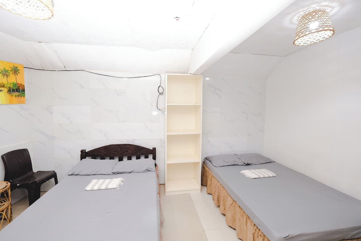 Una habitación pequeña y luminosa con dos camas individuales, un armario de madera, una silla de mimbre y una mesita.