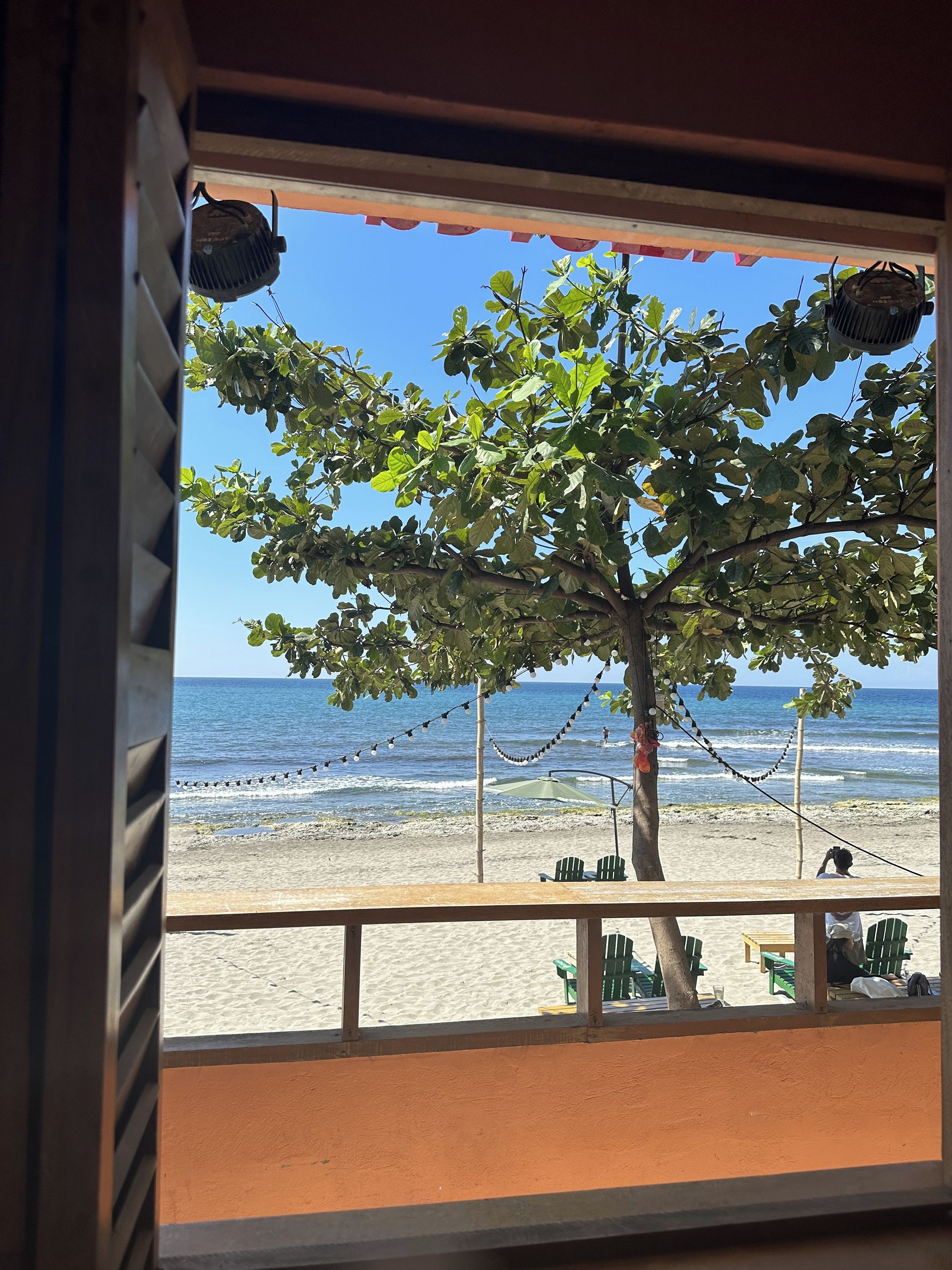 A beach view from a window with a tree, beach chairs, and a table.