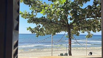 A beach view from a window with a tree, beach chairs, and a table.