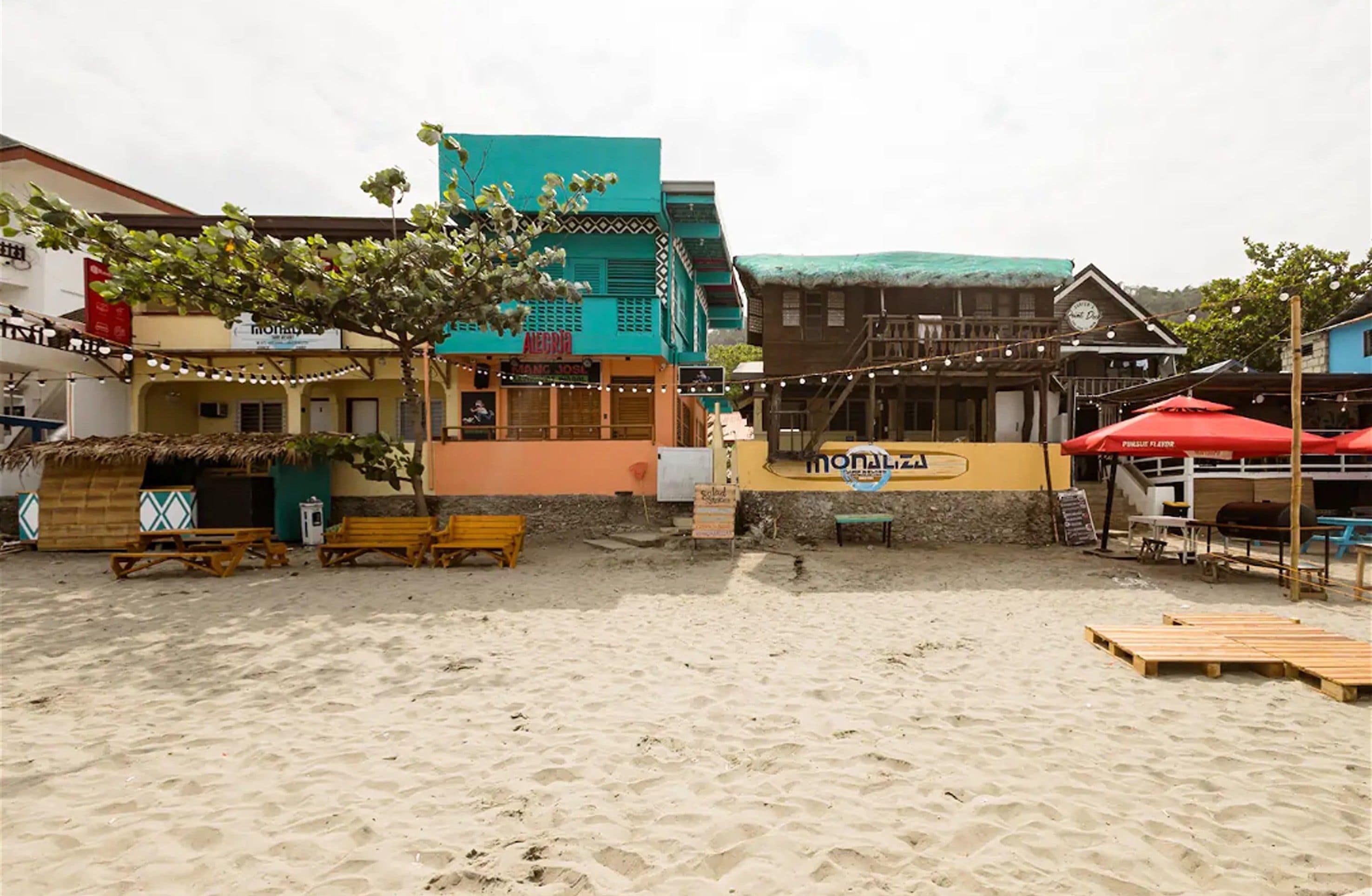 Una zona junto a la playa con sillones de madera, un cartel que dice 'Monaliza' y edificios con fachadas coloridas.