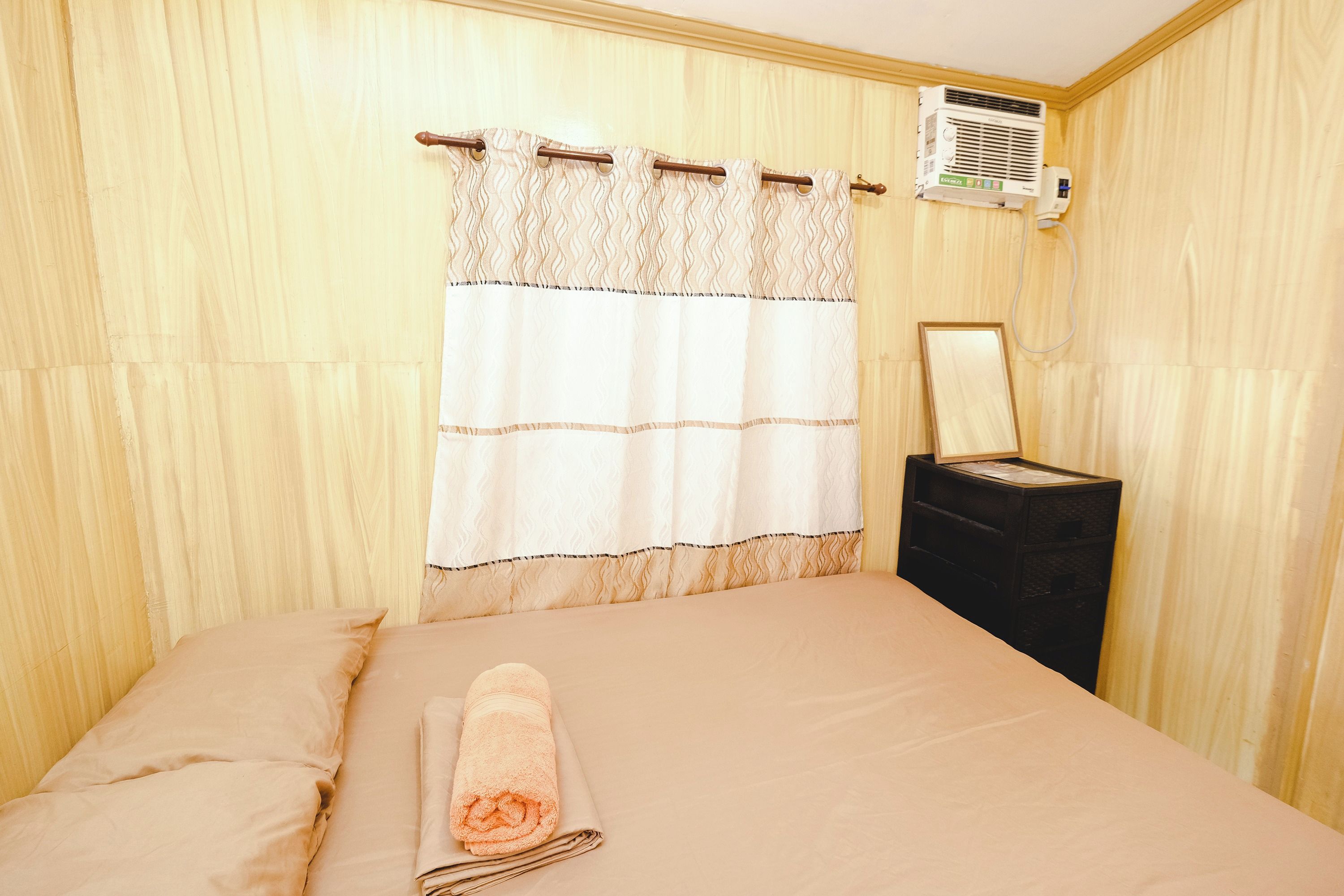 A hotel room with a bed, a rolled towel, a nightstand, a mirror, and a wall-mounted air conditioner.