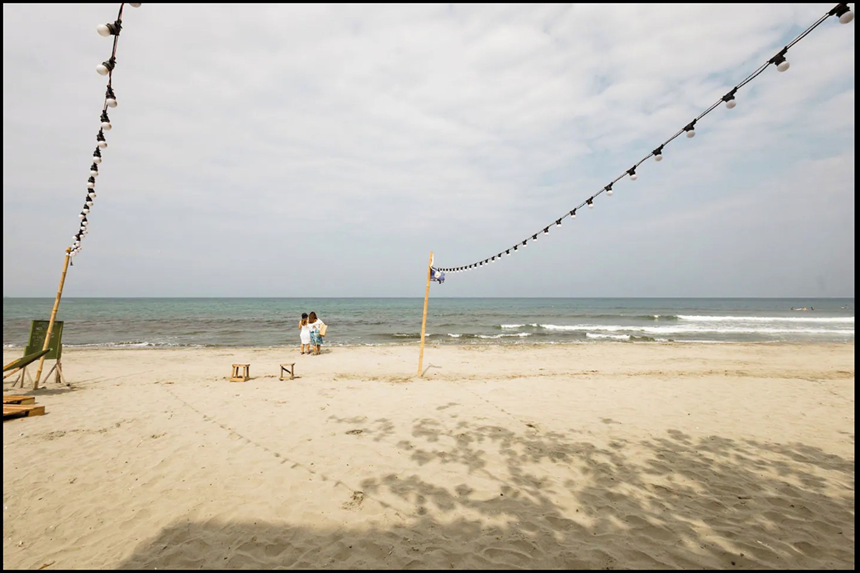 A beach with a row of lights, a wooden chair, and a couple sitting on the sand.