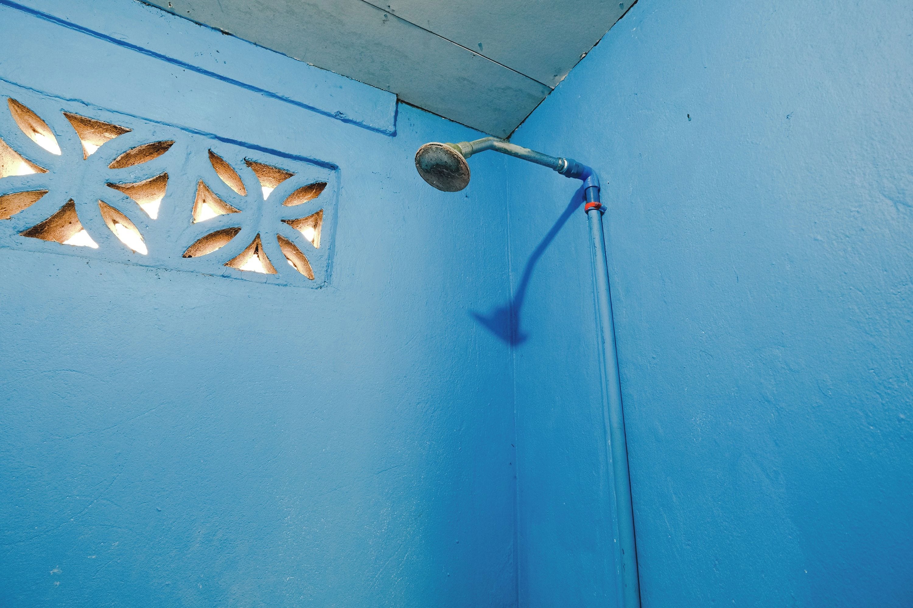 Un mur bleu avec une pomme de douche et une fenêtre ornée d’un motif géométrique.