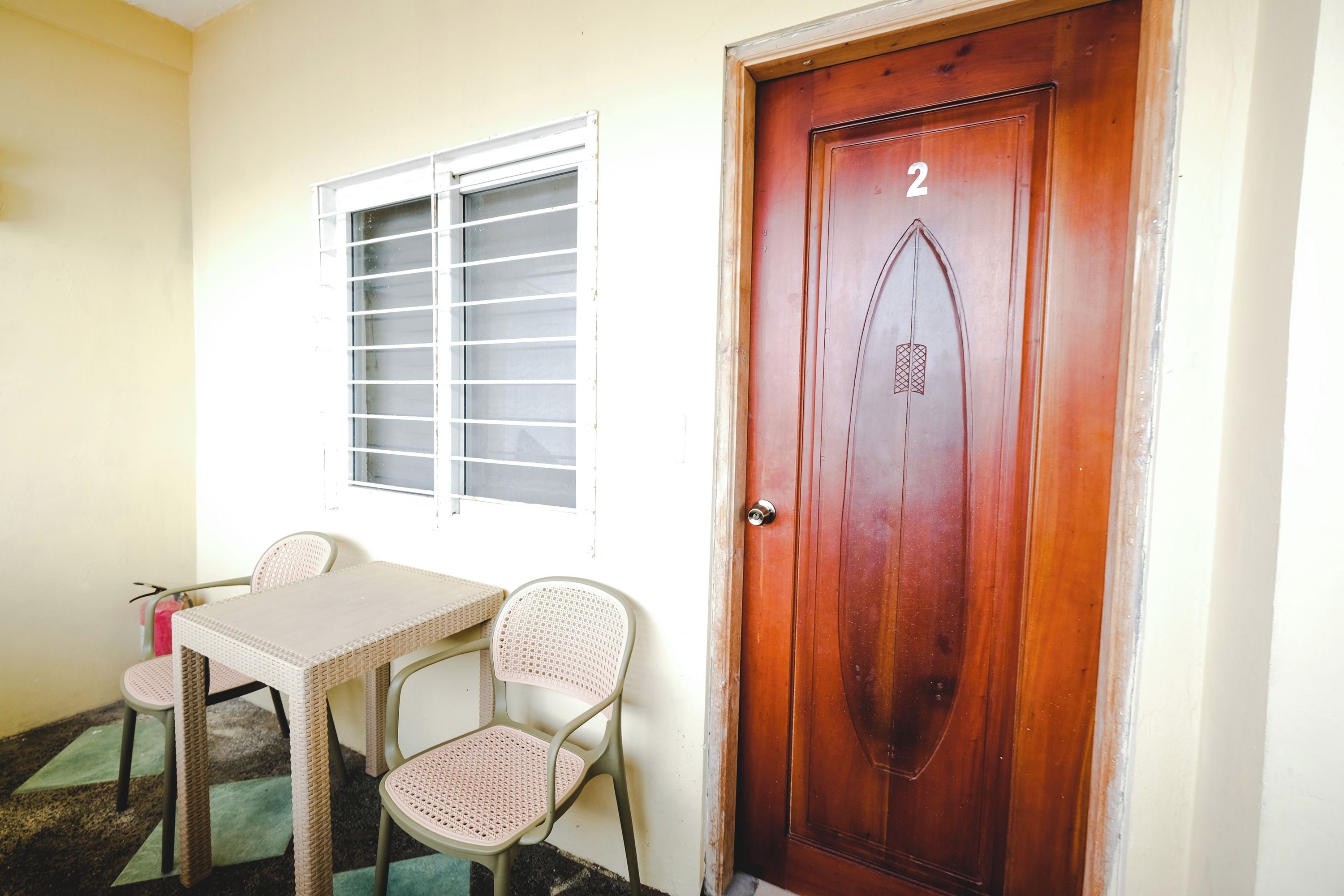 A room with a wooden door marked with the number 2, a window with metal bars, a wicker table, and two wicker chairs.