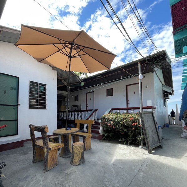 An outdoor seating area with a table and chairs under a large umbrella, a staircase leading to a second floor, and a balcony with a view of the sky. - Monaliza Surf Resort By Zuzu (San Juan)