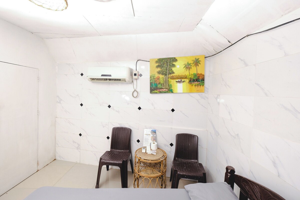 A hotel room with a bed, two wicker chairs, a small round table with a bottle and a tissue box, an air conditioning unit, and a painting of a landscape on the wall.