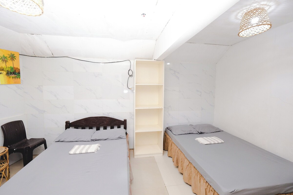 A small, well-lit room with two single beds, a wooden wardrobe, a wicker chair, and a small table.