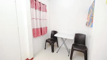 Una habitación pequeña con una mesa blanca y dos sillas de mimbre, una cortina roja y blanca, y un cuadro de colores en la pared.