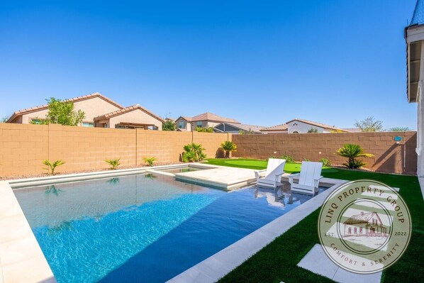 A swimming pool with a clear blue water surface, surrounded by a brick wall and a grassy area with two white lounge chairs.