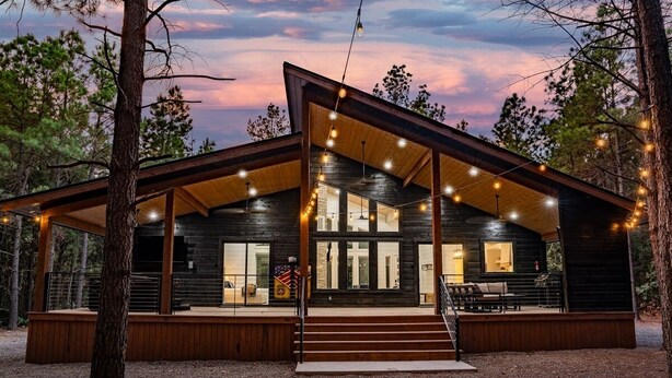 Une cabane en rondins moderne, dotée d’une terrasse en bois, de grandes fenêtres et de guirlandes lumineuses.