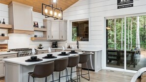 Une cuisine moderne avec des meubles blancs, un îlot central doté de tabourets, un luminaire suspendu et une vue sur l’extérieur grâce à une porte-fenêtre coulissante.