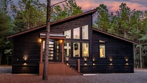 Une cabane en rondins avec une façade en bois, de grandes fenêtres, un escalier menant à l’entrée et une allée gravillonnée.
