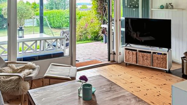 Un salon doté d’un parquet, d’un téléviseur posé sur un meuble, d’un canapé, d’une table basse et offrant une vue sur un jardin grâce à de grandes fenêtres.