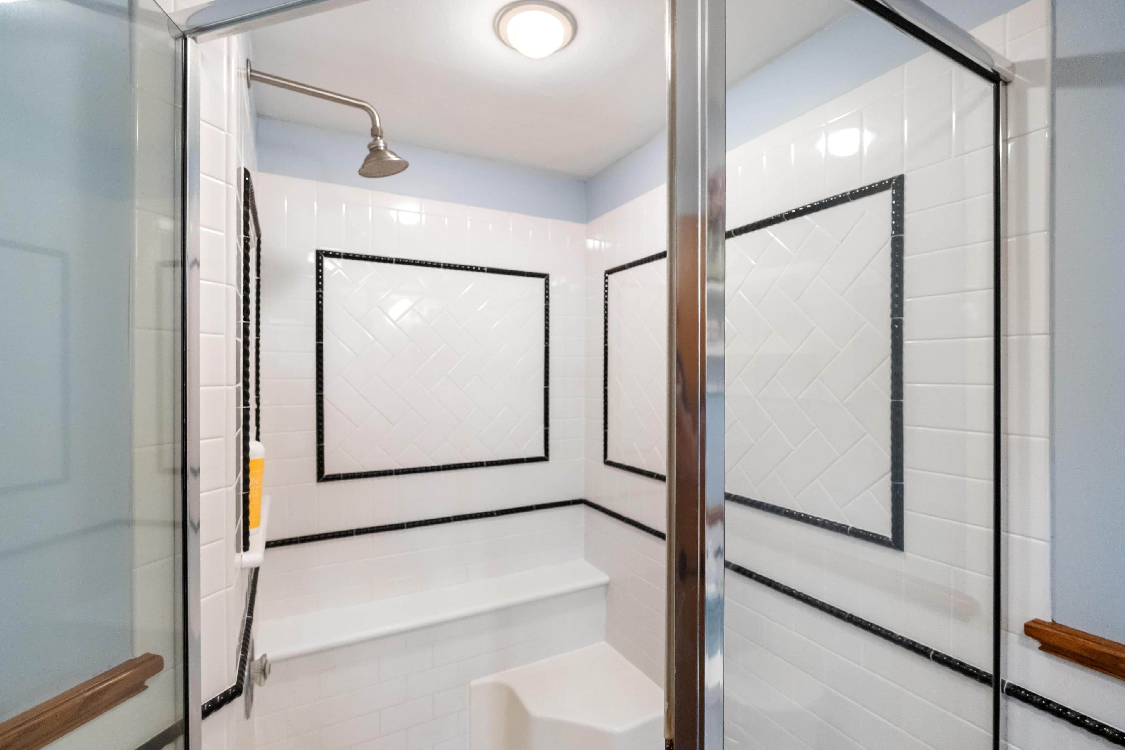 A glass-enclosed shower with a handheld showerhead, white tiled walls, and a small shelf.
