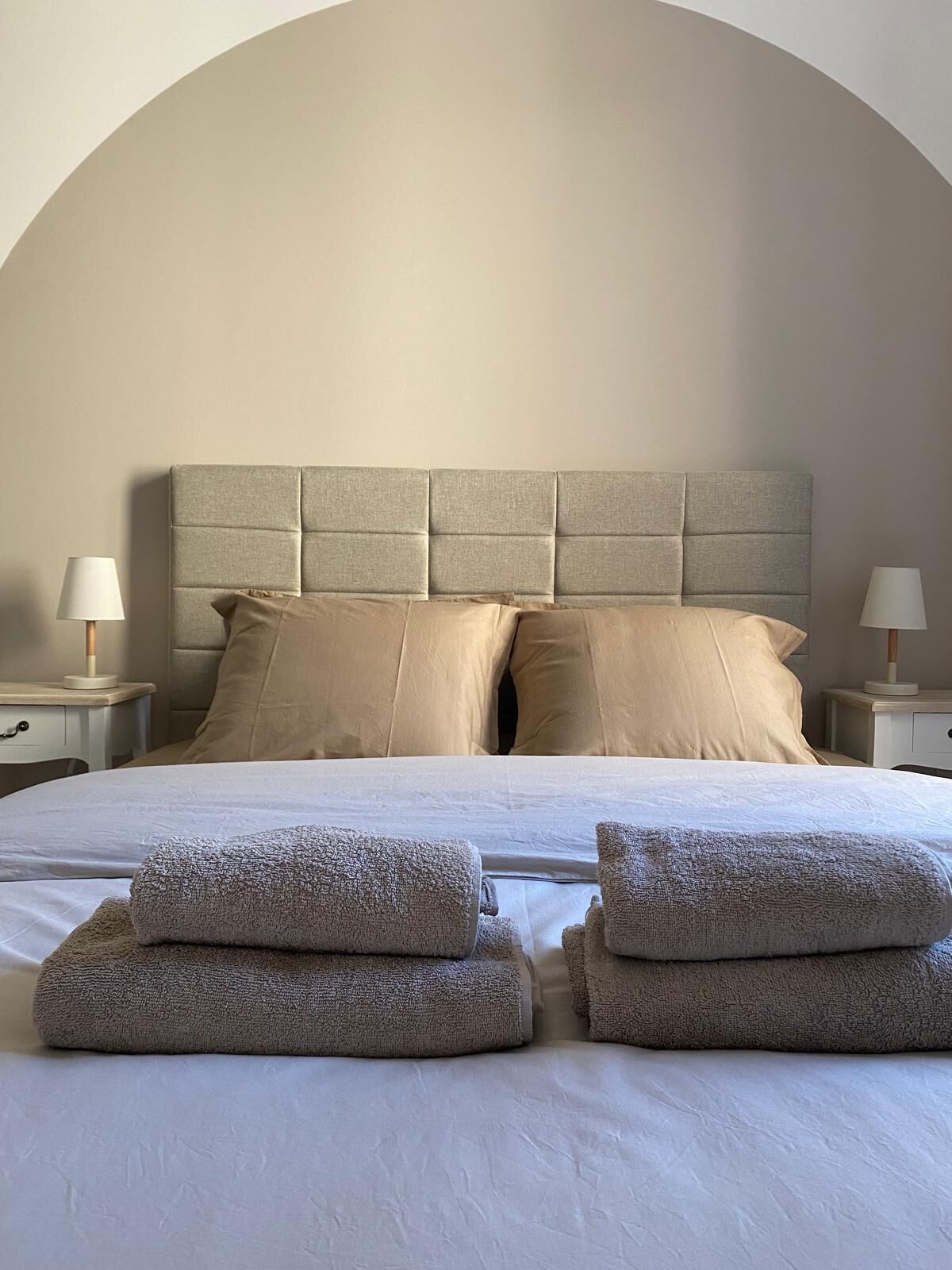 Una cama bien tendida con un cabecero capitonado, dos mesitas de noche con lámparas y un amplio ventanal en forma de arco sobre la cama.