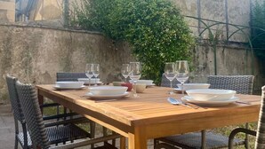 Un espace de restauration en plein air avec une table en bois et des chaises en osier, adossé à un mur de pierre orné de plantes grimpantes.