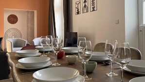 Un coin repas avec une table en bois dressée pour six personnes, entourée de chaises. Trois œuvres d’art encadrées ornent le mur, ainsi qu’un téléviseur et une porte avec des rideaux.