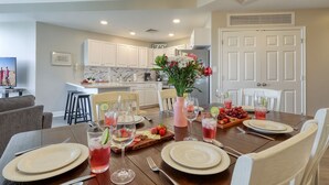 Un comedor con una mesa de madera dispuesta para cuatro personas, rodeada de sillas blancas. Se observa una cocina al fondo con armarios y una puerta, ambos en color blanco.