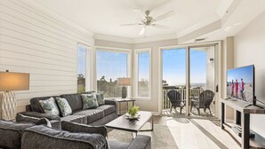 Amplia sala de estar con un sofá gris, una mesa de centro y un televisor sobre un mueble. Hay un balcón con vistas a la playa.