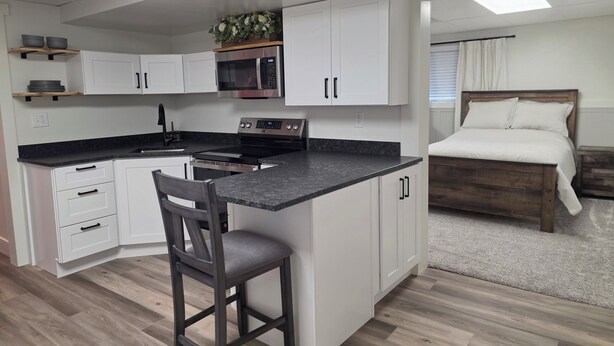 Une cuisine compacte avec des meubles blancs, un four à micro-ondes et une cuisinière. La pièce adjacente contient une chaise en bois et un lit avec une tête de lit en bois.