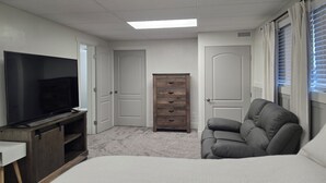 Une chambre d’hôtel dotée d’une grande télévision à écran plat, d’une commode en bois, d’un canapé gris et d’un luminaire au plafond.